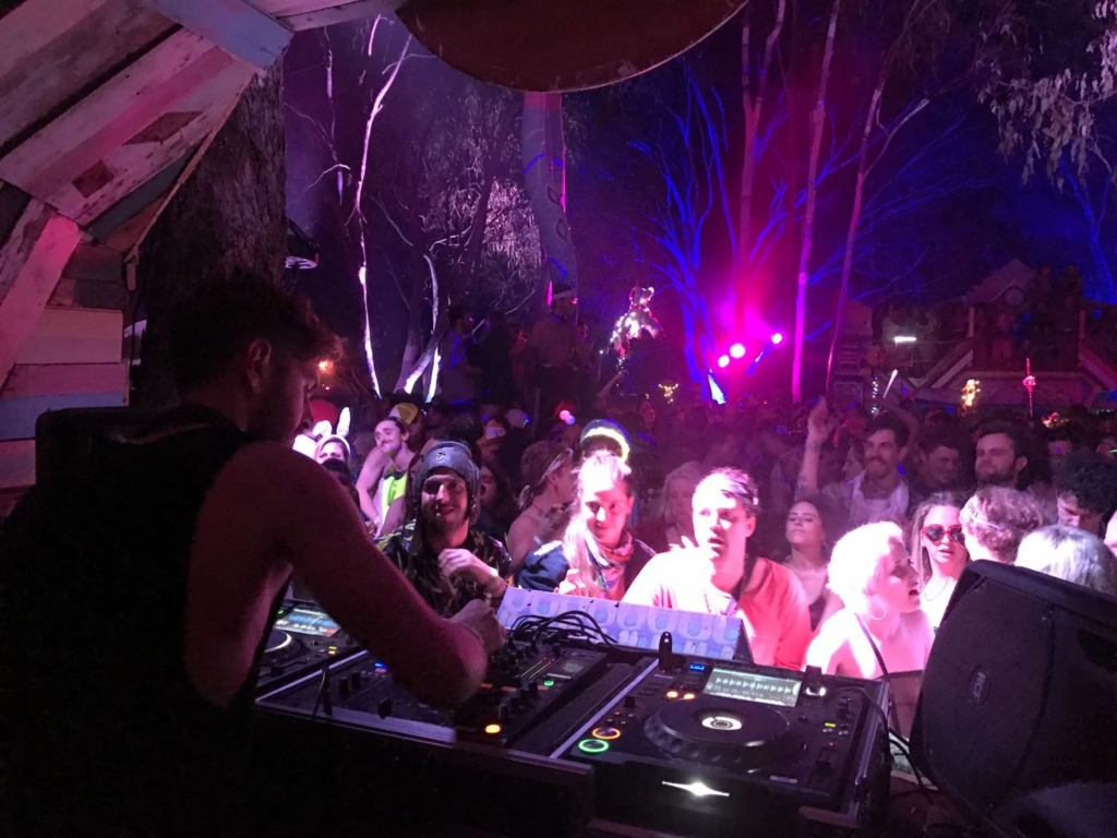 DJ Jozef Conor performs to a packed outdoor crowd at night, with colourful stage lighting and festival dancers near the booth.