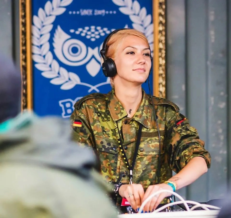 Deysa performs at an event wearing headphones and a camouflage jacket, standing behind the DJ booth in front of a blue venue backdrop.