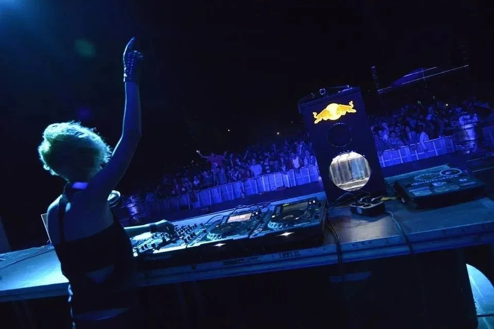 Romanian Techno DJ and producer Deysa performs at a night festival, raising one arm from behind the decks in front of a large crowd under blue stage lighting.