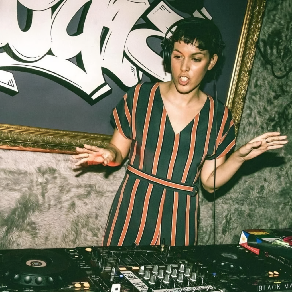 Mica performs behind the decks with headphones on, hands raised over the mixer during a live DJ set.