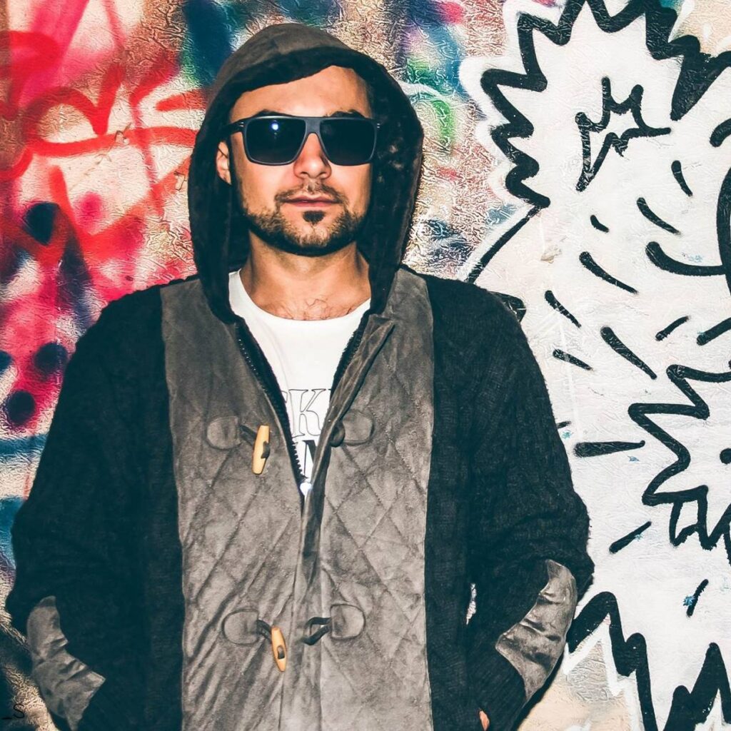 Samotarev stands in front of a graffiti wall wearing sunglasses and a hooded jacket.