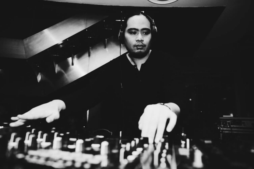 Black-and-white photo of DJ Mike Reefer performing behind the decks with headphones on during a live DJ set.
