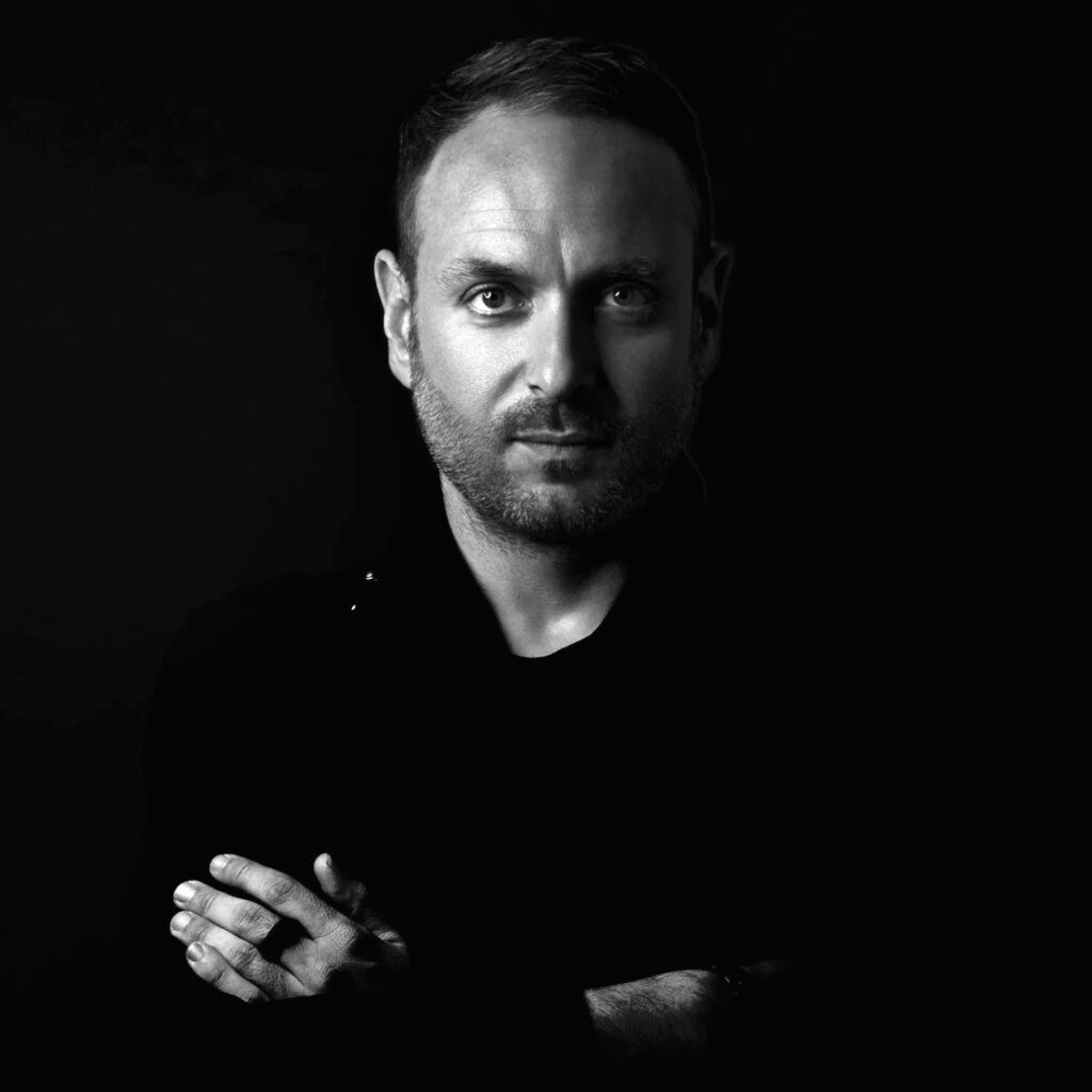 Black-and-white portrait of DJ Ruby standing with his arms crossed against a dark background.