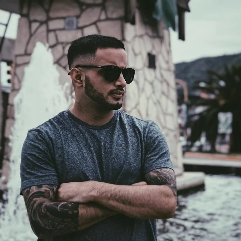 Lio Q wearing sunglasses and a grey T-shirt with folded arms, standing beside a fountain and looking to the left.