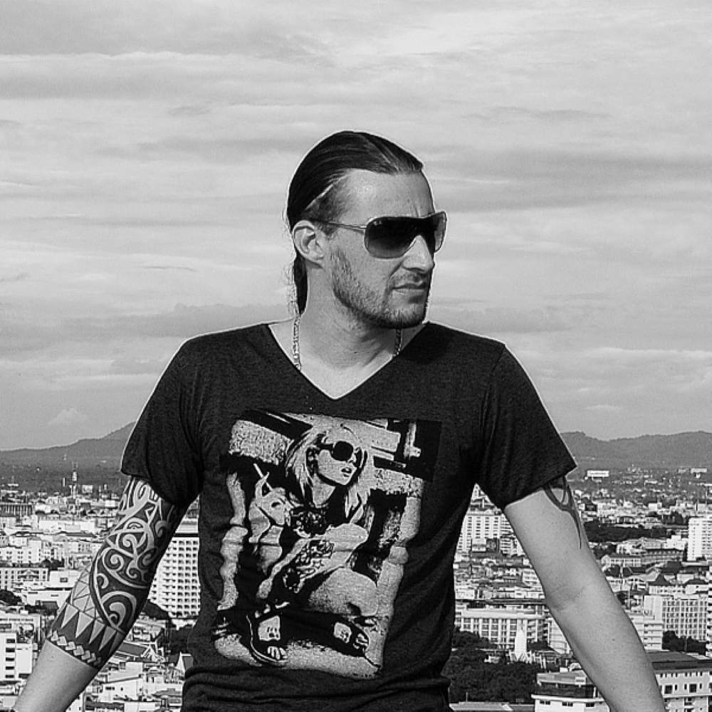 Black-and-white portrait of PatriZe wearing sunglasses and a graphic T-shirt while standing on a rooftop overlooking a city.