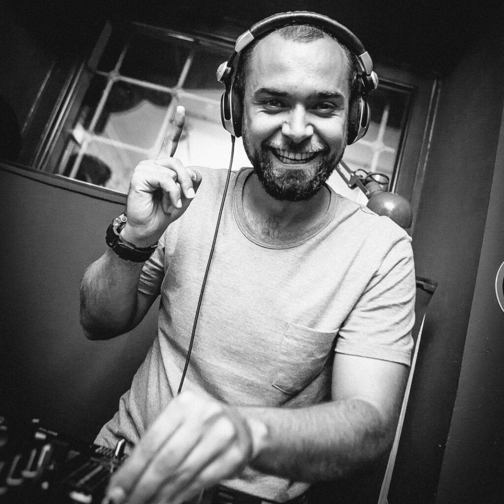 Black-and-white image of Samotarev smiling while performing in headphones, with one finger raised above the DJ equipment.