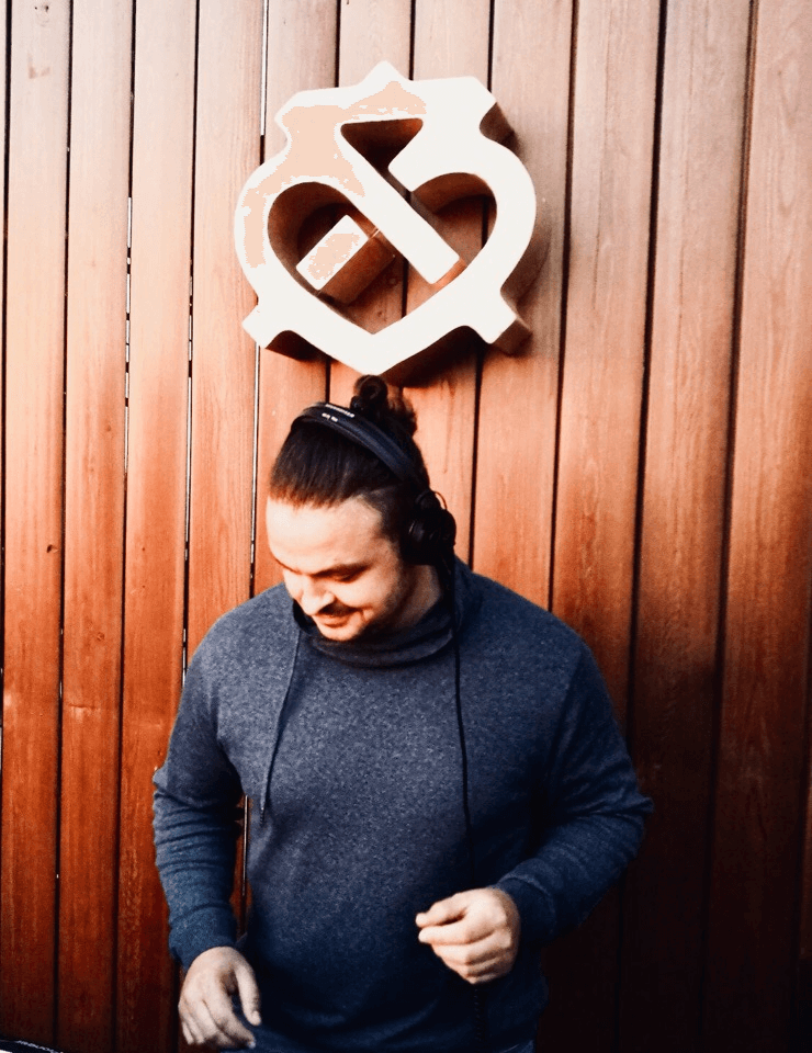 Bolgarin wearing headphones and looking down while performing in front of a wooden wall with a white logo above him.
