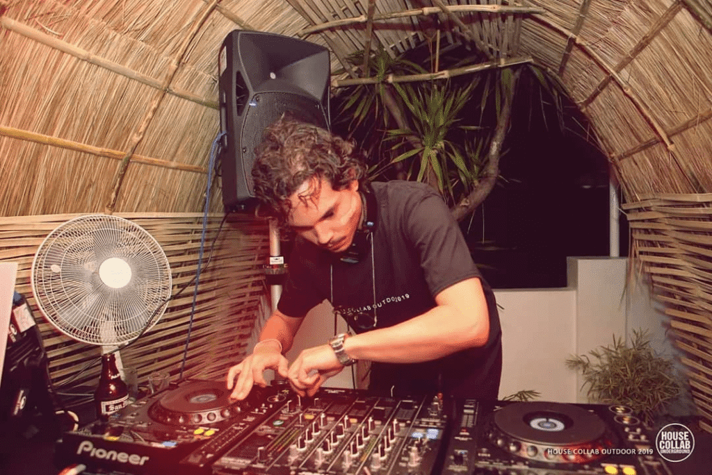 DJ Adri Malaki performing on Pioneer equipment beneath a thatched roof at an outdoor night event.