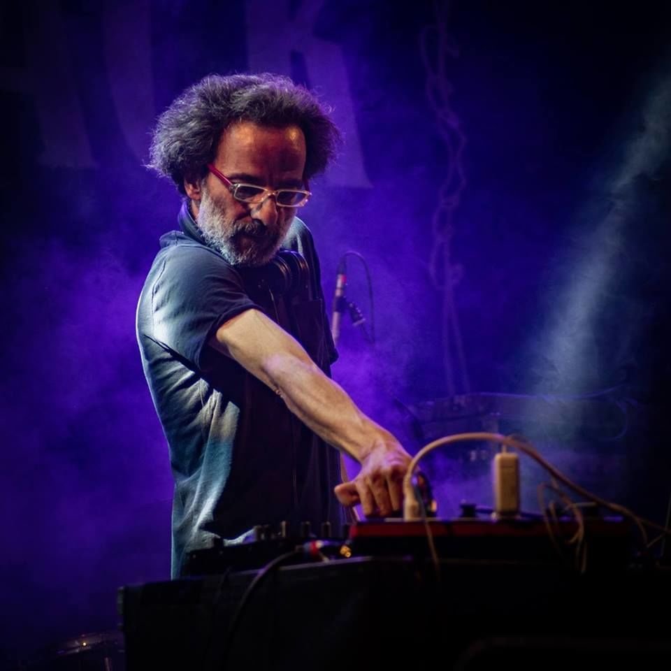 Roby M Rage performing on DJ equipment under purple stage lighting and smoke, with headphones around his neck.
