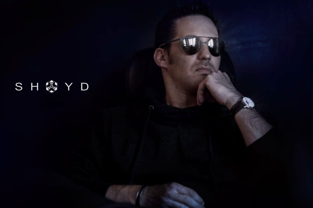 Shoyd sitting in sunglasses with one hand resting against his chin, against a dark blue background with his name displayed beside him.