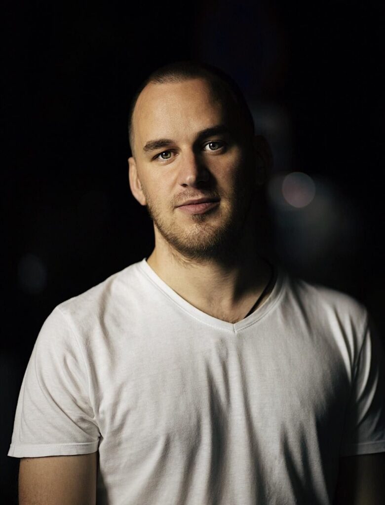 Studio portrait of Ilya Gerus wearing a white T-shirt against a dark background.