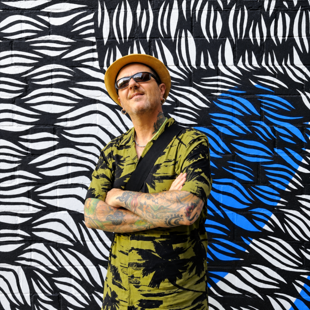 Italian DJ and producer El Brujo wearing a straw hat, sunglasses and a green patterned shirt, standing with his tattooed arms crossed in front of a black, white and blue graffiti wall.