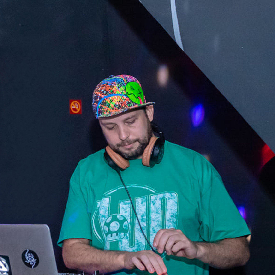 Yushka, Nottingham-based DJ and promoter, performing at Broma with a laptop, wearing a colourful splatter-print cap and headphones around his neck.