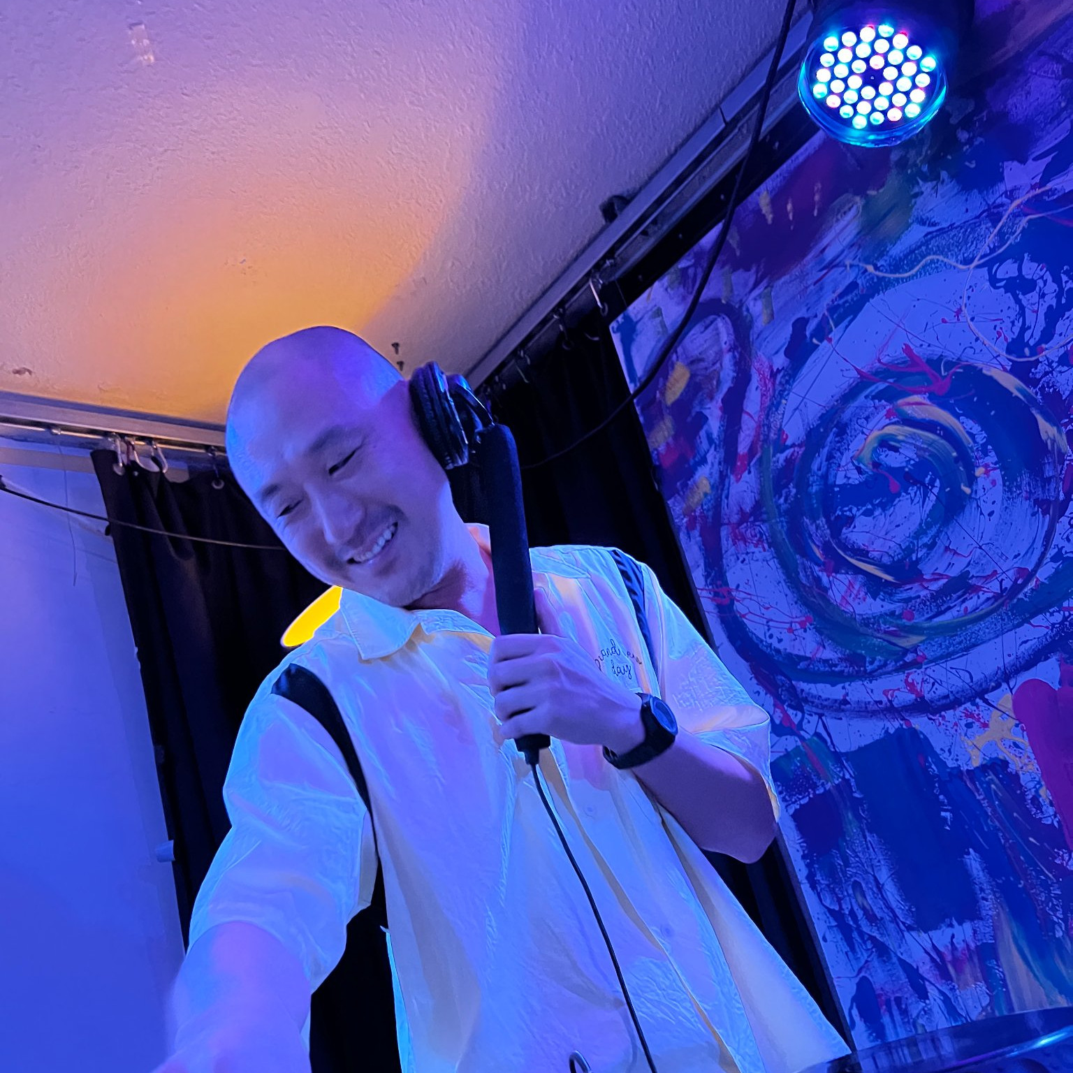 Taiwanese DJ Murphy Kin performing at an event holding the headphone to his left ear with very blue l;ighting, colour image.