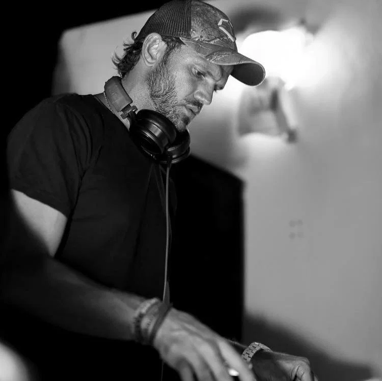 Black-and-white image of Poli Siufi wearing a cap and headphones around his neck while performing at DJ equipment, representing the Argentine DJ and producer originally from San Salvador de Jujuy.