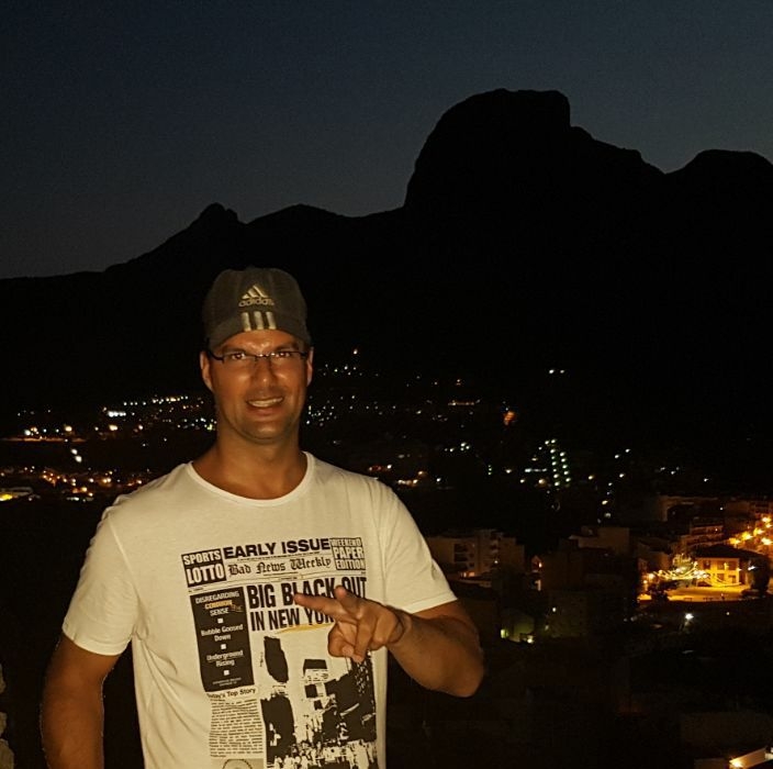 Dutch DJ and producer Basscontroll standing on a hill above a town at nightimes wearing a baseball cap, colour image.