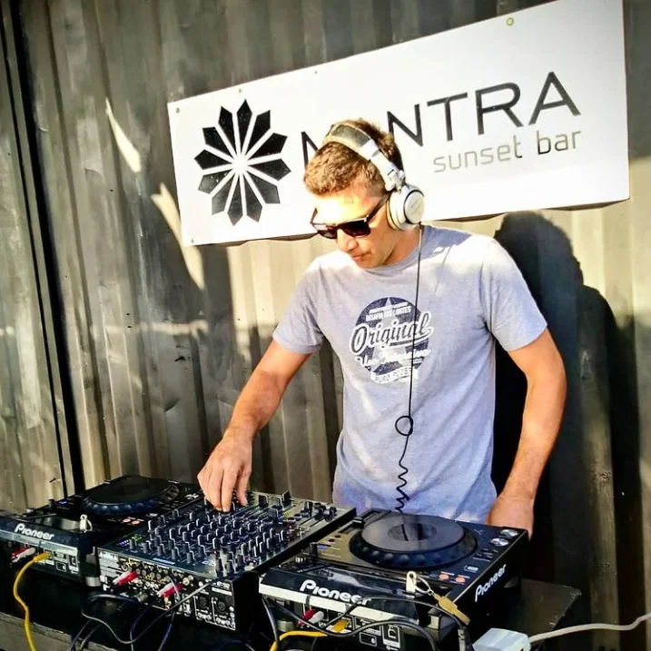 Deep Juss performing on Pioneer CDJs and mixer at Mantra Sunset Bar, headphones around his neck and sunglasses on.
