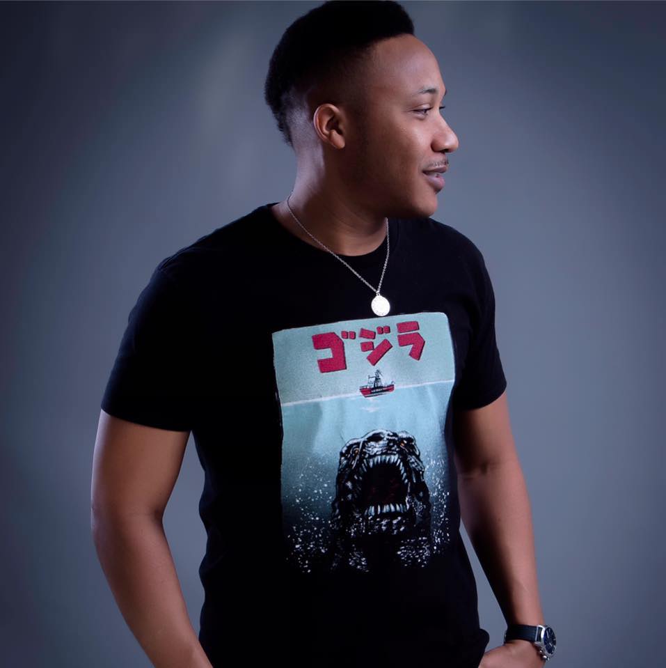 DJ Gerard Jones wearing a black T‑shirt with a Godzilla design, silver necklace, and wristwatch, standing against a neutral gray background, facing to the right.