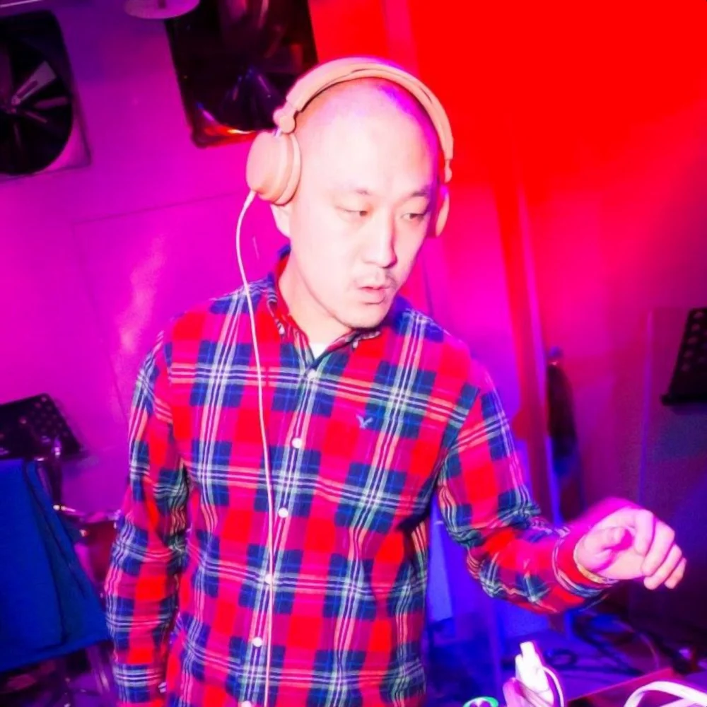 Taiwanese DJ and promoter Murphy Kin performing as a DJ wearing pink headphones and a check shirt, colour image.