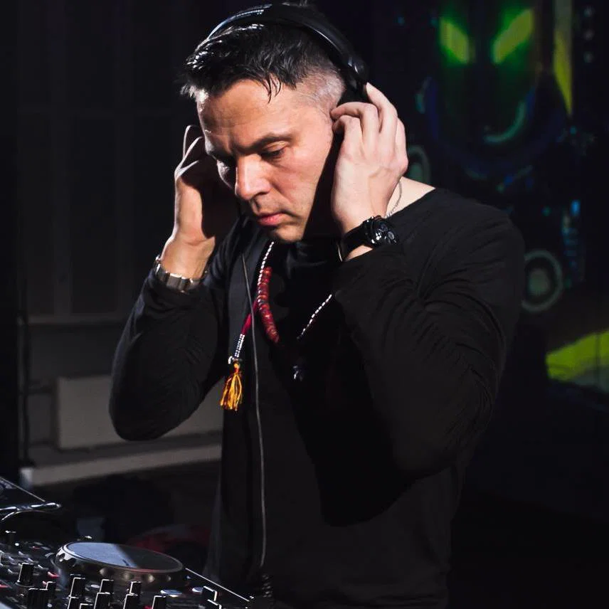 K Loveski cueing a track at the decks, hands on headphones, against a dark club backdrop with colourful UV artwork.