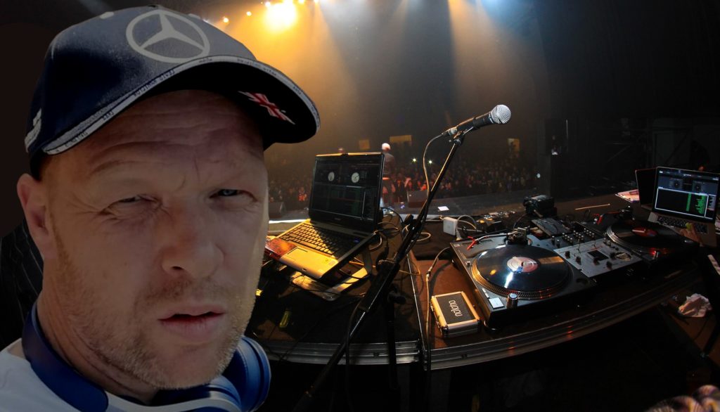 DJ Bean There performing at an event with crowd and turntables behind him looking at camera, colour image.