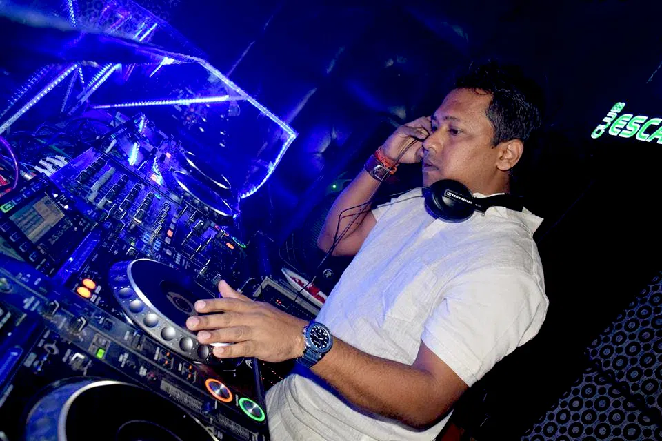 Indian DJ and producer Psy Inertia wearing headphones and looking down at the DJ equipment whilst performing at an event, taken from his left side in a colour image.