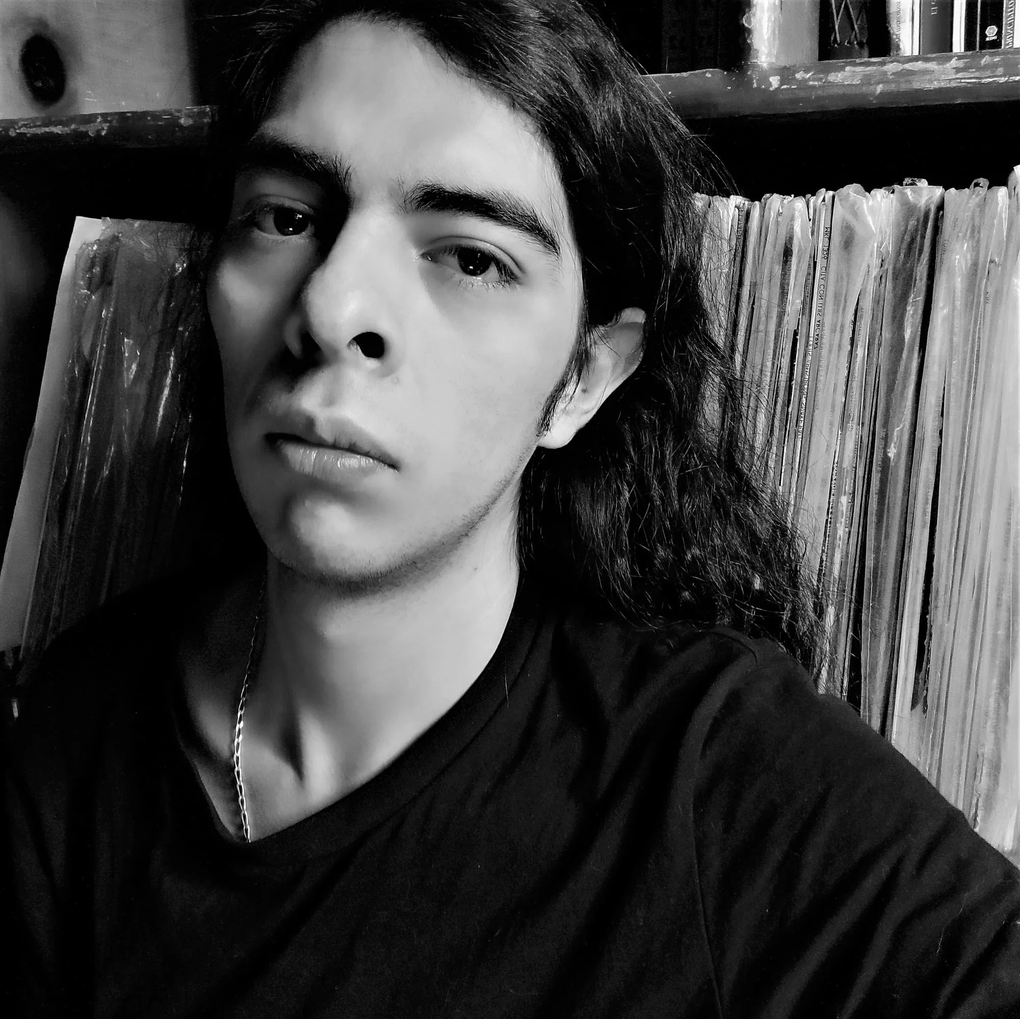 Black-and-white portrait of electronic music artist Danz Maxine seated beside a collection of vinyl records, wearing a dark shirt and looking directly at the camera.