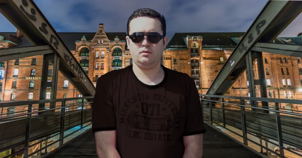 Portuguese DJ Joshua standing in front of a building on a bridge over a river at dusk, colour image.