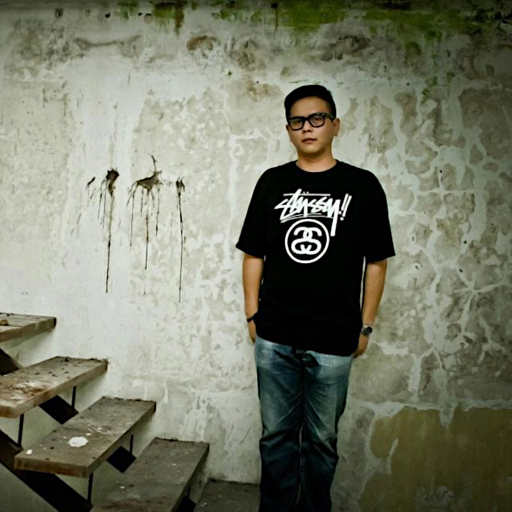Malaysian DJ and producer Terence C standing with his hands by his side looking at camera in front of dirty wall with wooden stairs to his right, colour image.