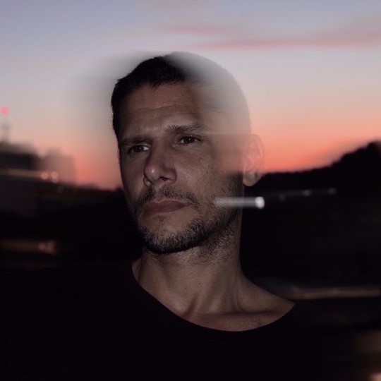 Brazilian DJ and producer Ricky Ryan standing outside at daybreak in a upper body shot with a time lapse picture, colour image.