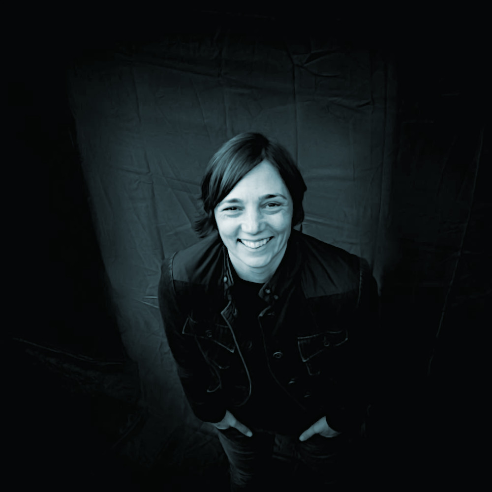Dutch DJ and producer Miss Dominguez tsnding and leaning forward with her hands in her pockets smiling at the camera, black and white image with blue tinge.