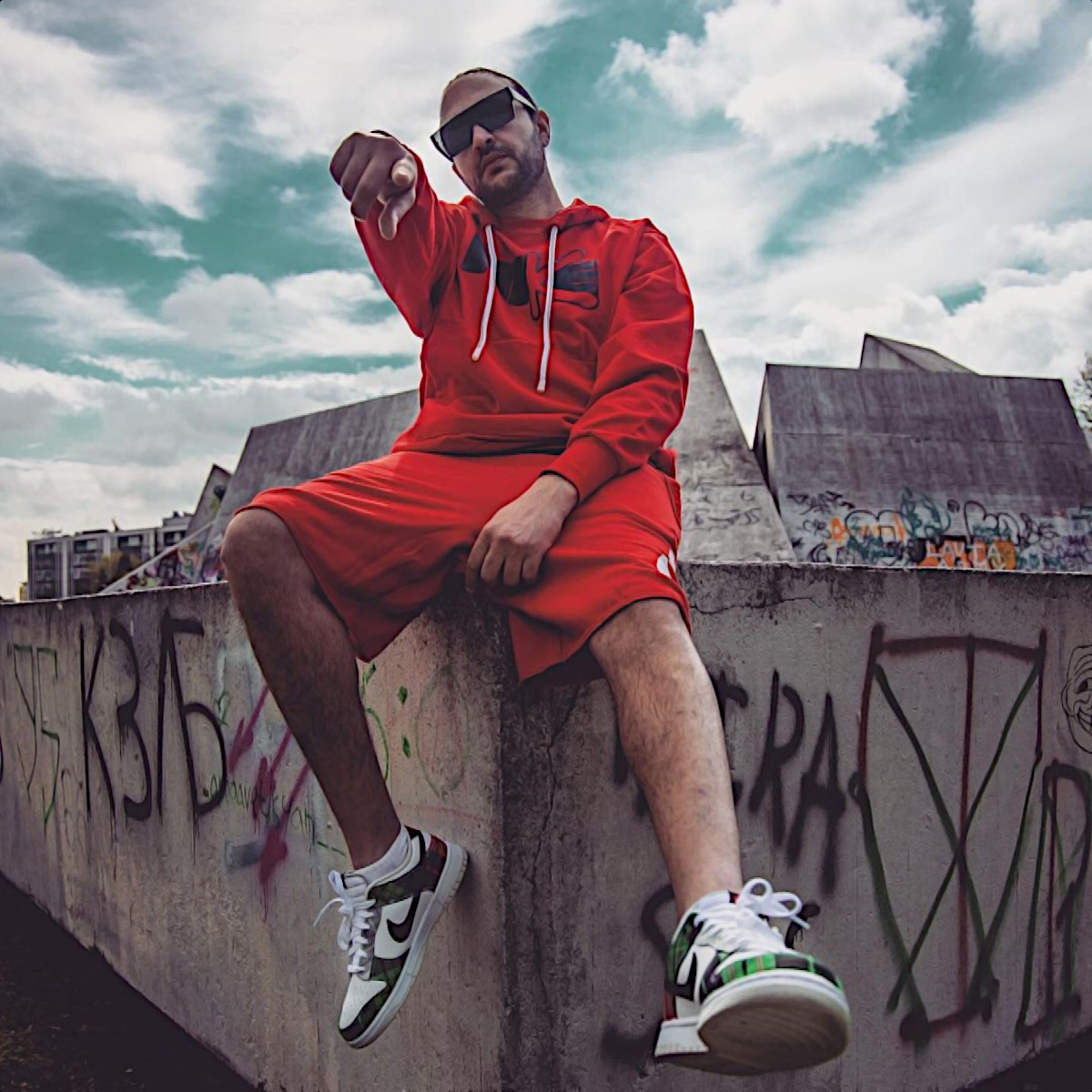 Bulgaria based DJ and producer ISMAIL.M sitting on the corner of a wall pointing his finger at thne camera wearing sunglasses and street wear, colour image.