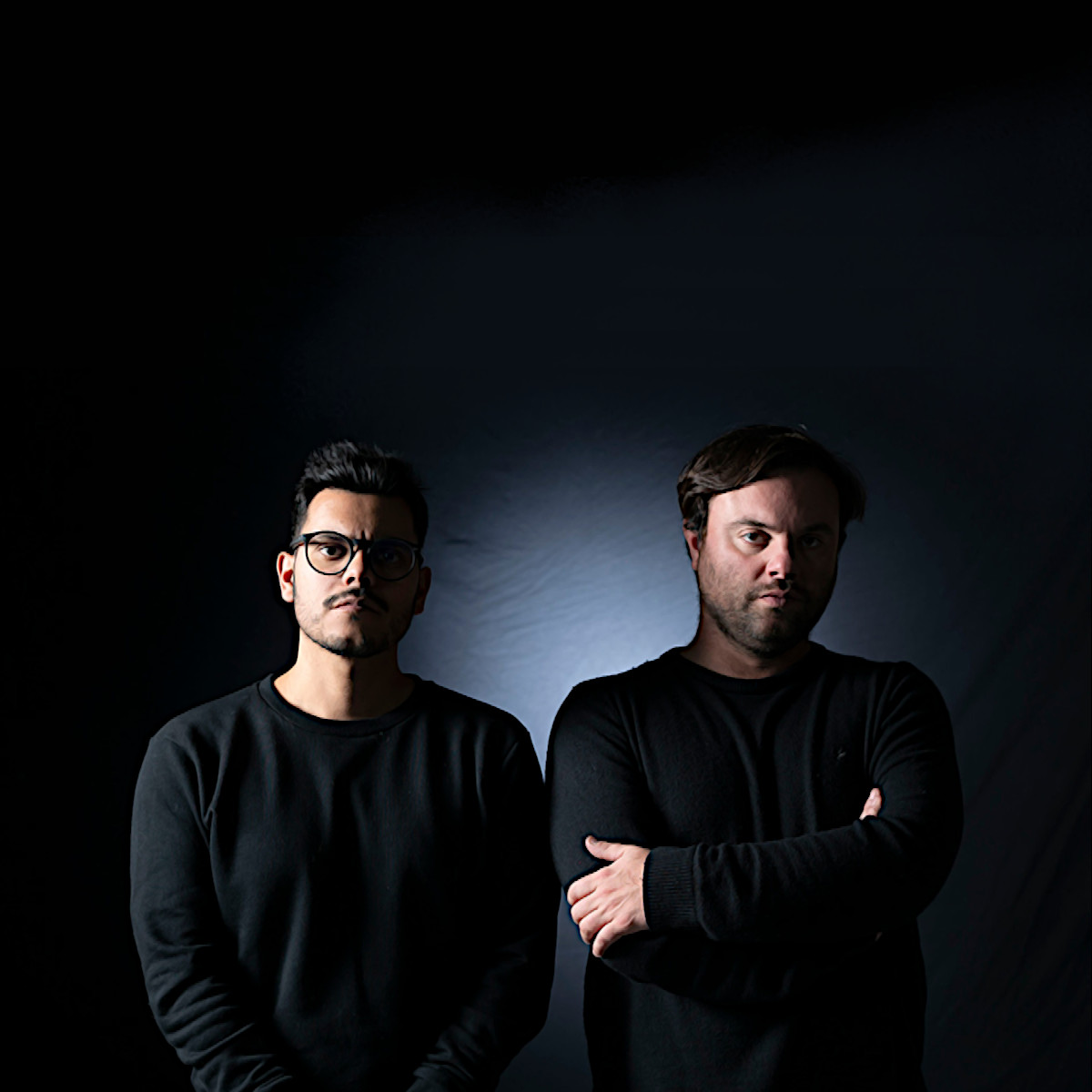 Argentinian DJs and producers Melodiam (AR) standing side by side on a black background with some lighting behind them, colour image.