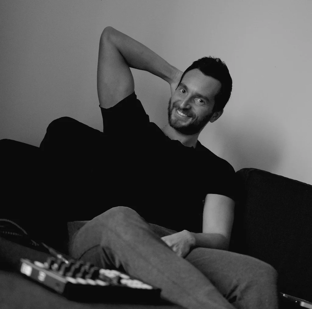 Italian DJ and producer Inve sitting on sofa with his right hand behind his head smiling, sampler on table in front of him, black and white image.