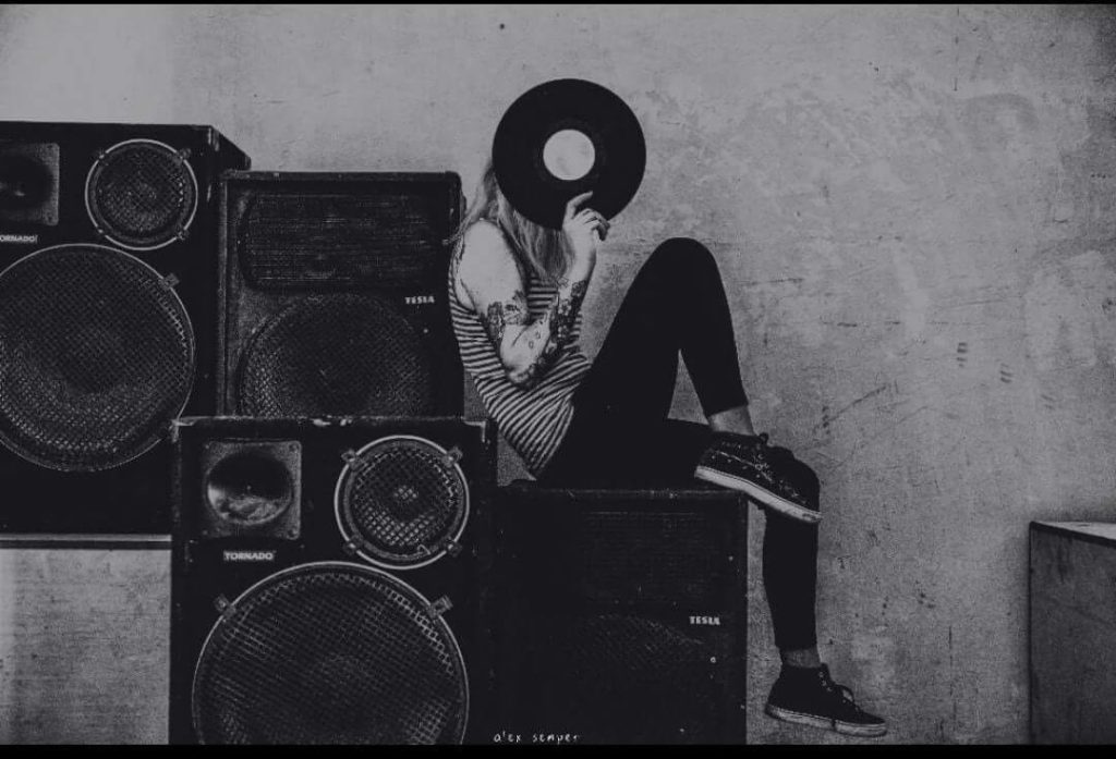 Romanian Techno DJ and Producer Deysa sitting sideways on Speakers and Blocking the side of here face with a vinyl record, black and white image.