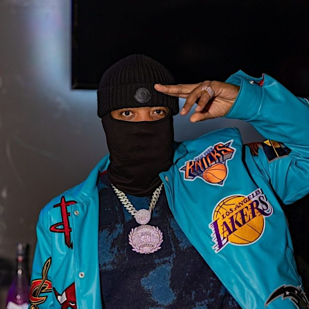 American Hip Hop Rapper Grea8Gawd standing wearing a face mask and doing a left handed two finger salute wearing an NBA jacket with several teams badges on it, colour image.