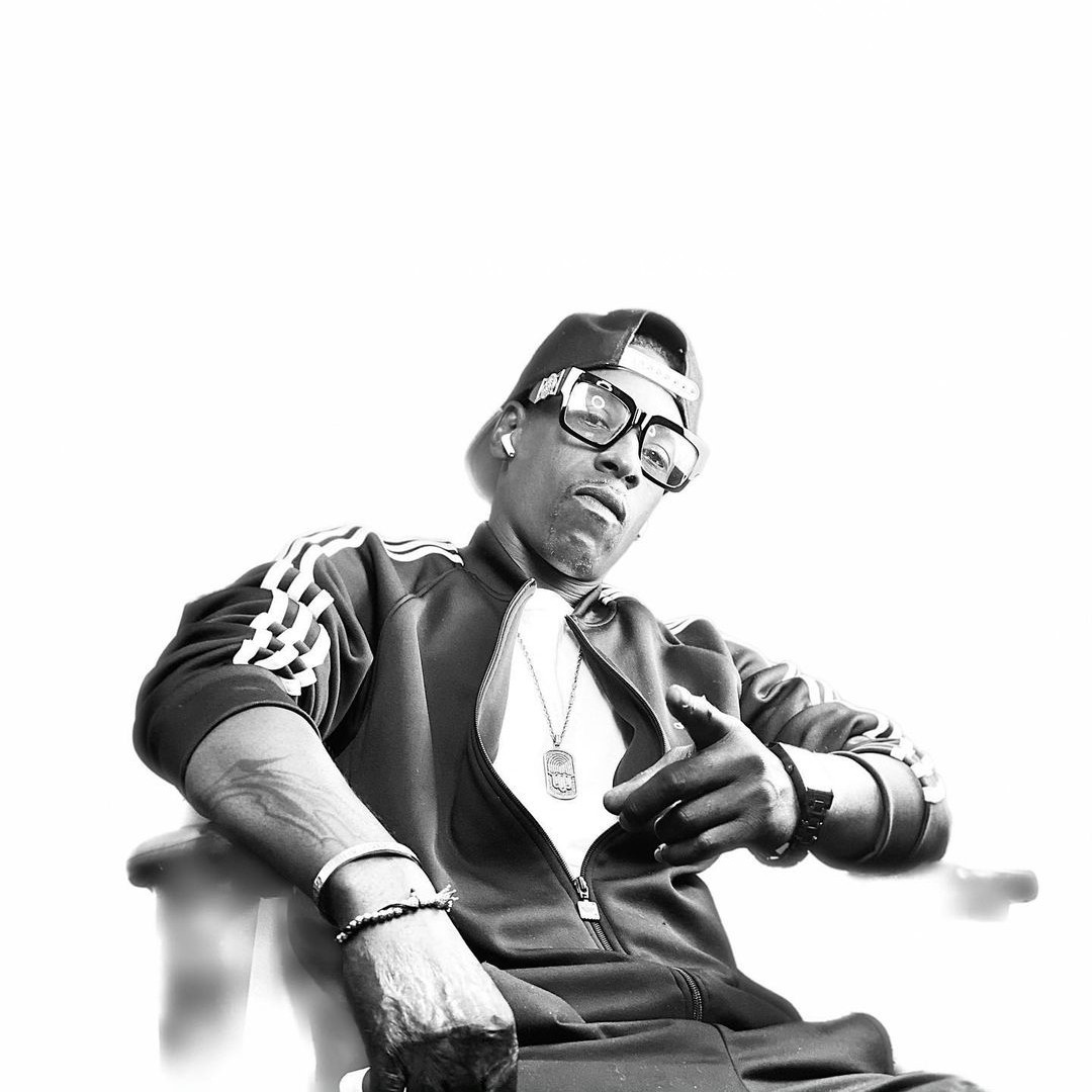American rapper and producer Imam T.H.U.G sitting in achair wering glasses and a baseball cap backwards and pointing with his left hand at the camera, black and white image.