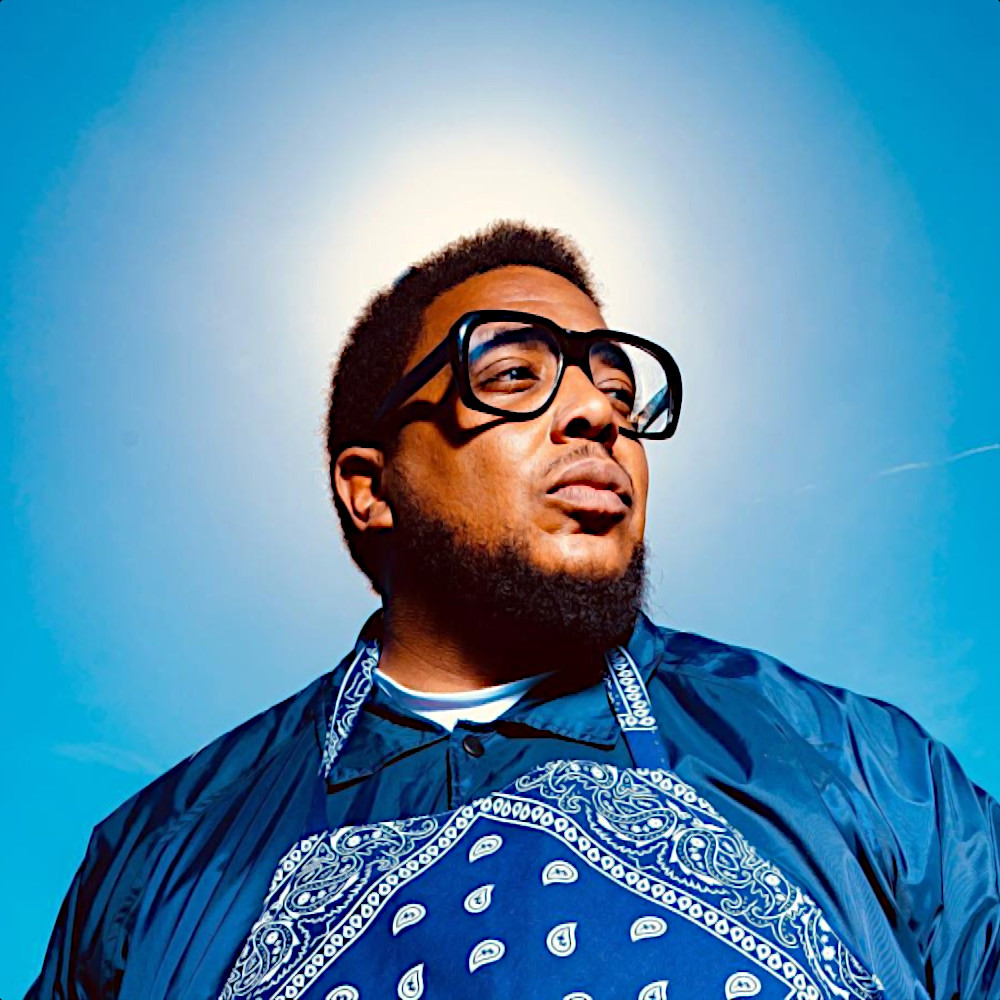 Nhà sản xuất và rapper người Mỹ Glasses Malone trong một bức ảnh chụp từ trên xuống ngoài trời, với bầu trời xanh làm nền; anh đeo kính gọng lớn trong suốt và đang nhìn sang bên trái, ảnh màu.
