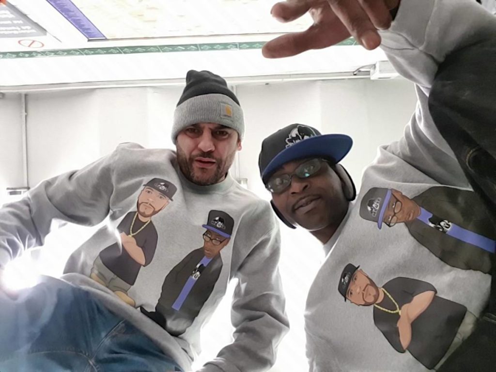 DJ Ronsha & G-Zon in a photo taken from below them lkooking down at camera wearing sweatshirts with cartoon images of themselves, colour image.