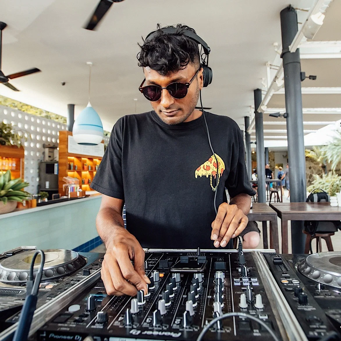 Sivanesh, Singapore-based house and techno DJ, performing on Pioneer DJ equipment at an open-air daytime bar venue wearing sunglasses and headphones.