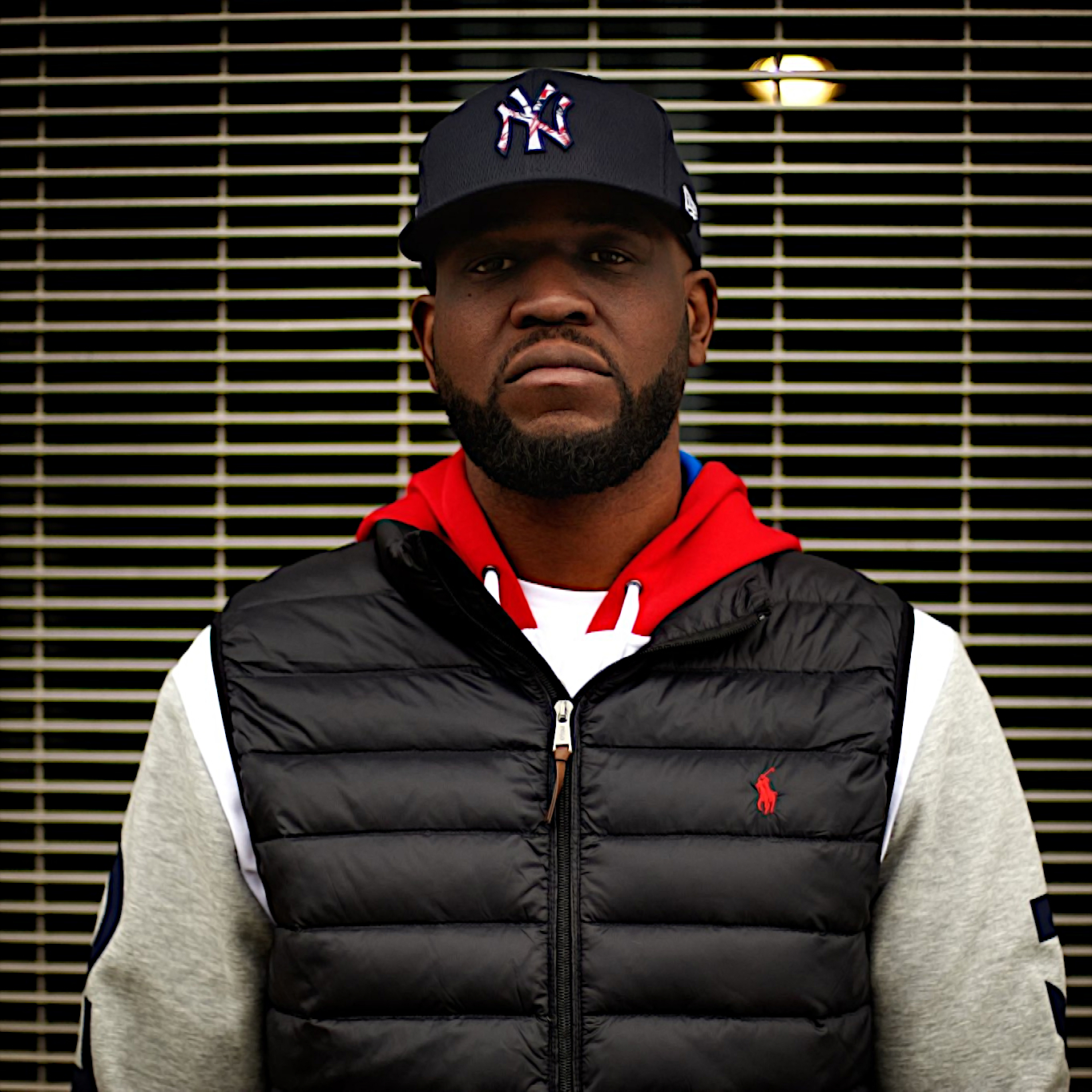 Canadian Rapper and producer Connaisseur Ticaso standing in upper body shot looking at camera wearing a baseball cap and sleeveless padded jacket over a hoodie, colour image.