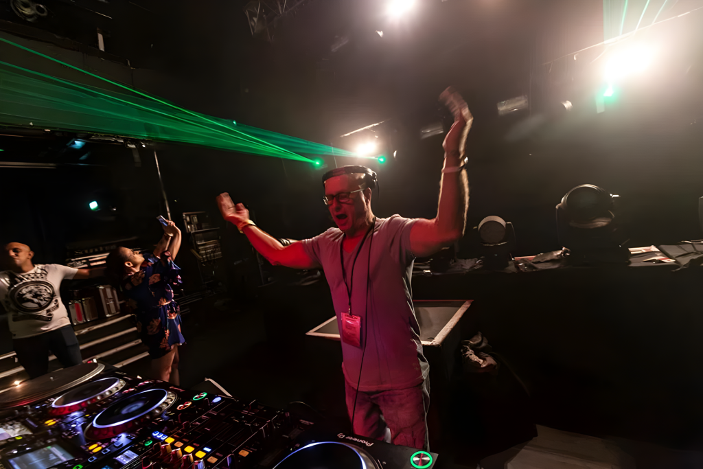 DJ Graham Gold raises his hands during a live club set, with Pioneer DJ equipment, stage lights and green lasers behind him.
