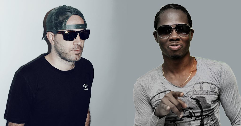 British DJ Dimensions wearing sunglasses and sunglasses and looking left standing next to Nigerian DJ Sly Fly also wearing Sunglasses and pointing at camera, colour image.