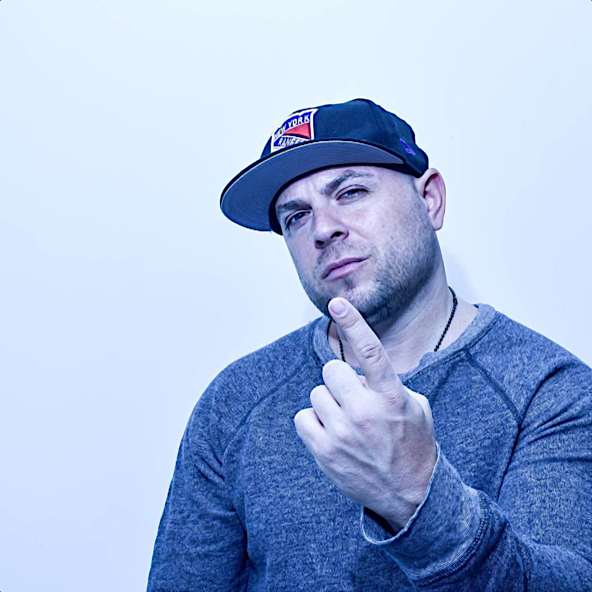 American Lyricist and rapper El Gant wearing a baseball cap with his right hand raised and his index finger pointing upwards, smiling wrly at the camera in an upper body shot, colour image.