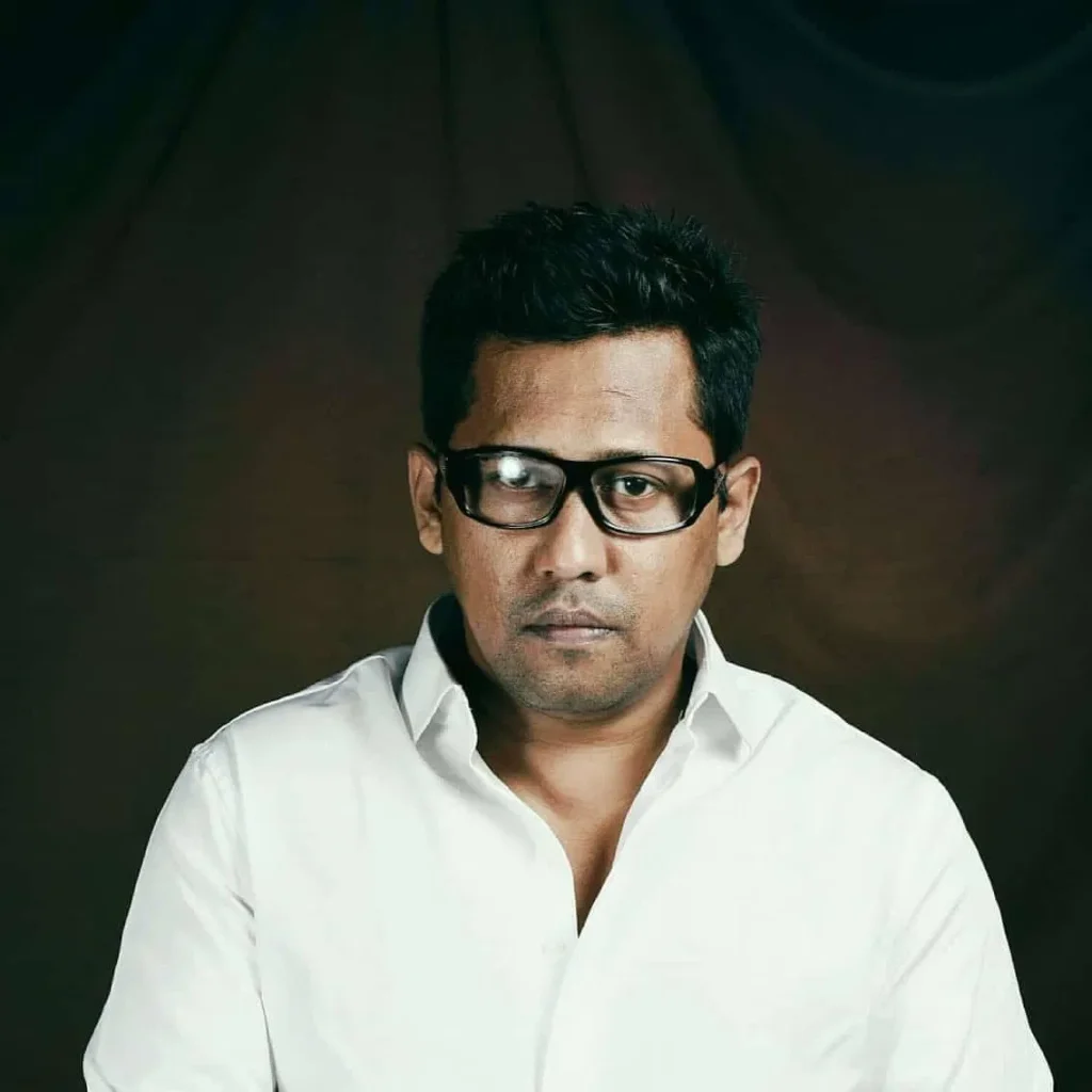 Indian DJ and producer Psy Inertia, also known as Kafka, in a studio portrait wearing black glasses and a white shirt against a dark background in a colour shot.