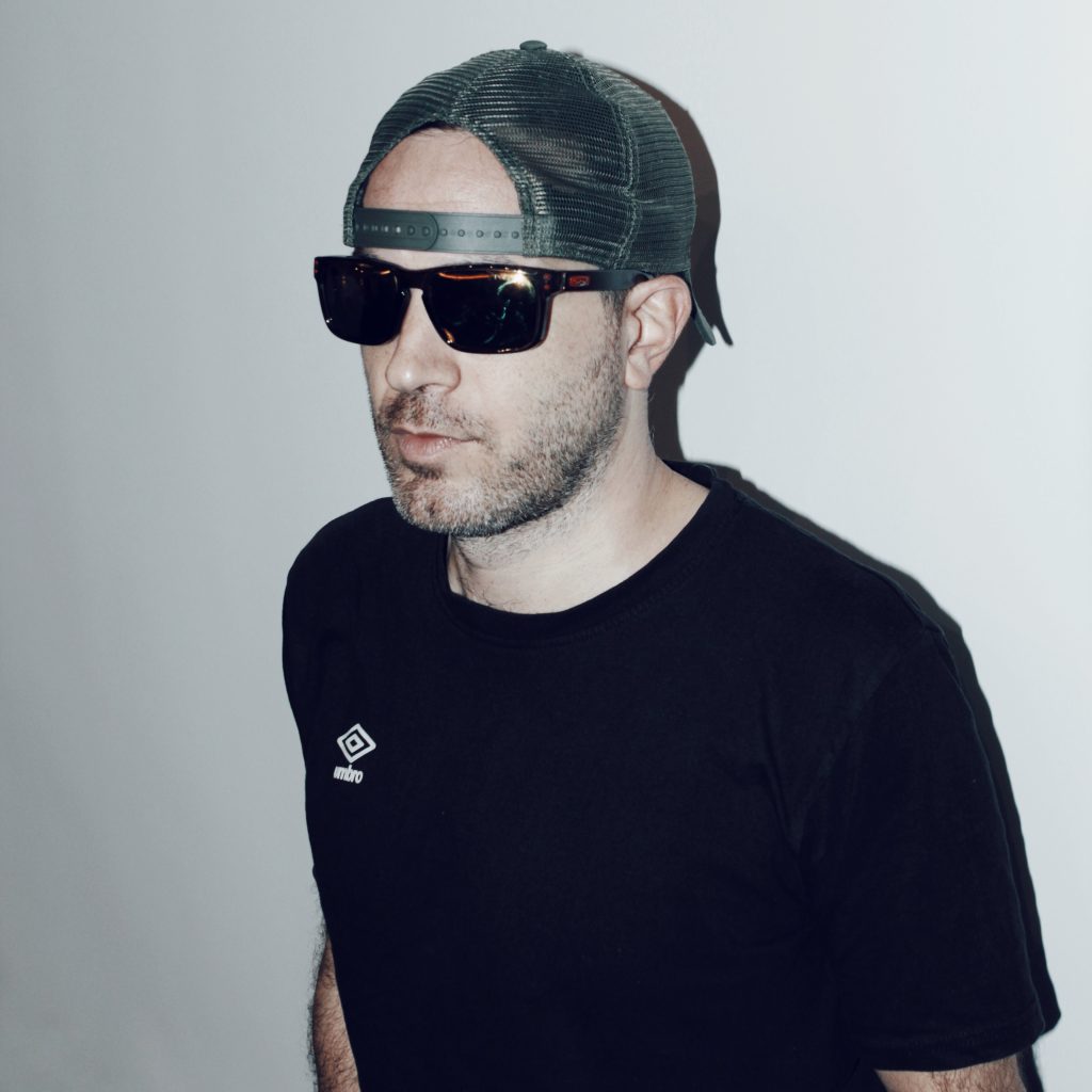 British DJ and promoter Dimensions standing looking to his right wearing sunglasses and a baseball cap, colour image.