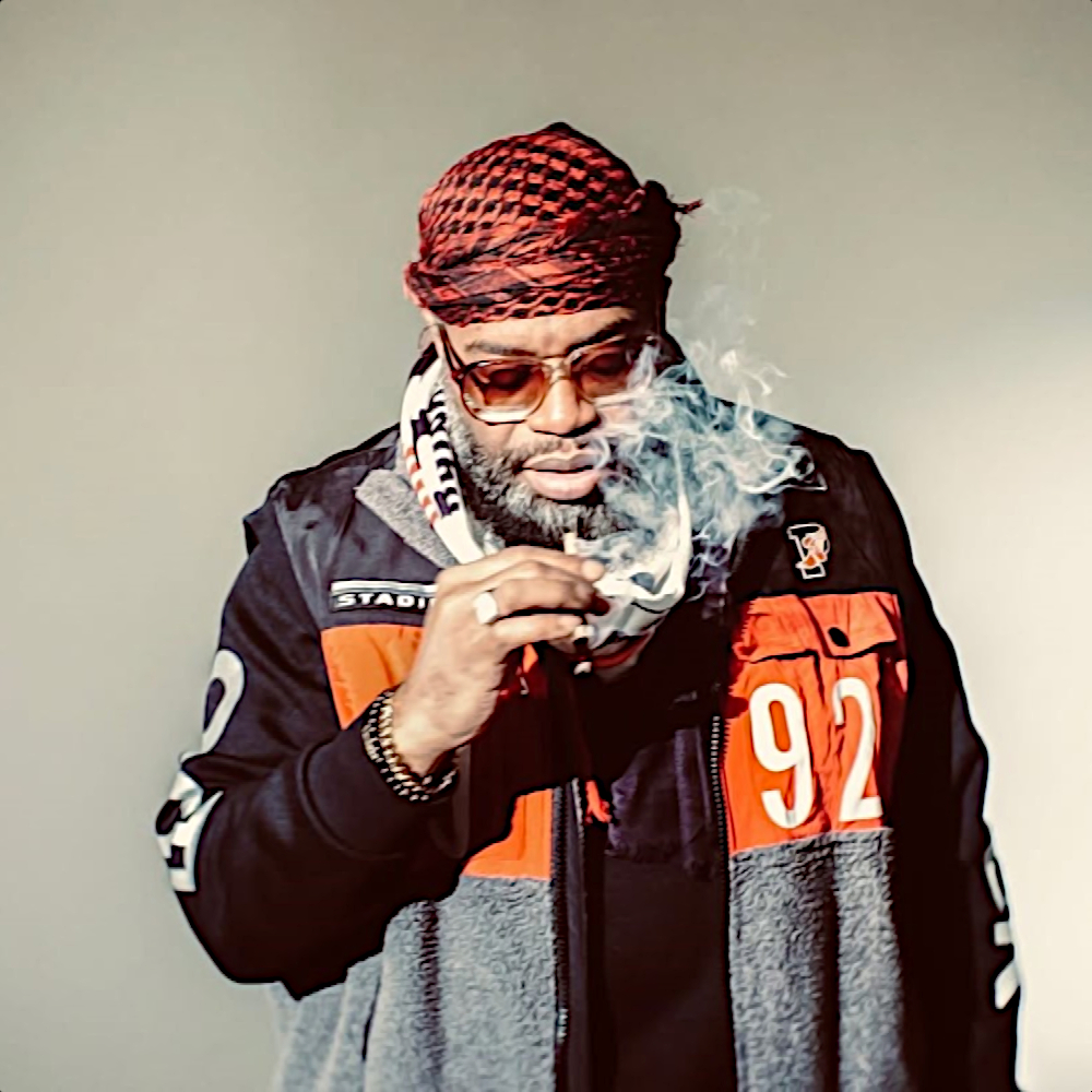 Ca sĩ rap và nhà sản xuất người Mỹ D.V Alias Khryst mặc áo khoác, đeo kính râm và quấn khăn shemagh, đang hút một điếu cần sa trong khi nhìn xuống đất, ảnh màu.