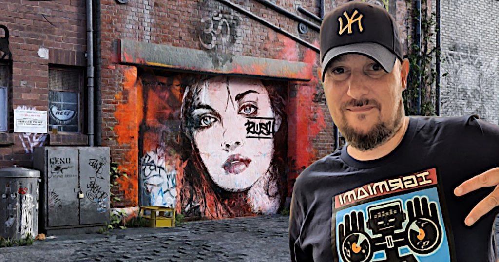 French Hip Hop DJ Phoenixx standing in front of a graffiti mural with a woman's face giving a two fingered gun salute, colour image.