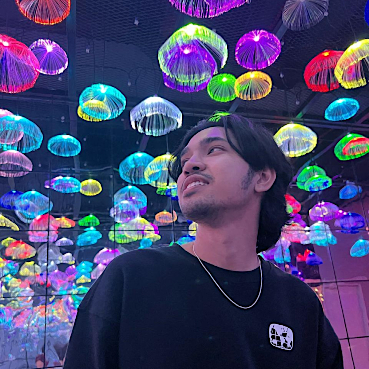 Malaysian DJ Teehee standing in a hall with many clear coloured umbrellas in the ceiling looking to his right and smiling, colour image.