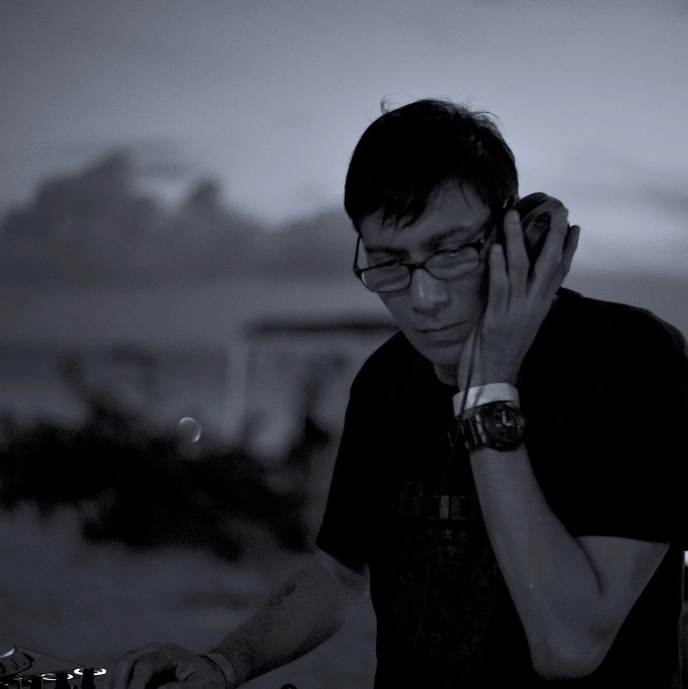 Black-and-white image of Brendon P holding one headphone while performing, representing his long-running work across Singapore’s club scene and international House music circuit.